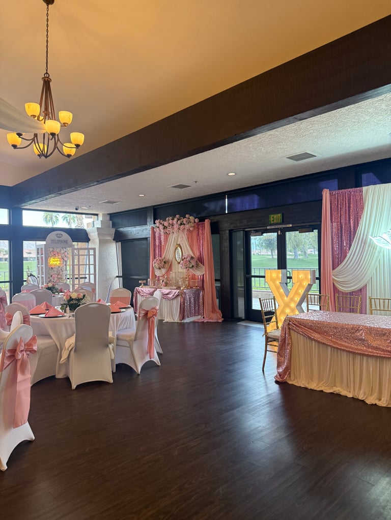 Elegant event venue decorated with pink and gold accents, round tables with chair covers, draped curtains, and ornate lighting fixtures.