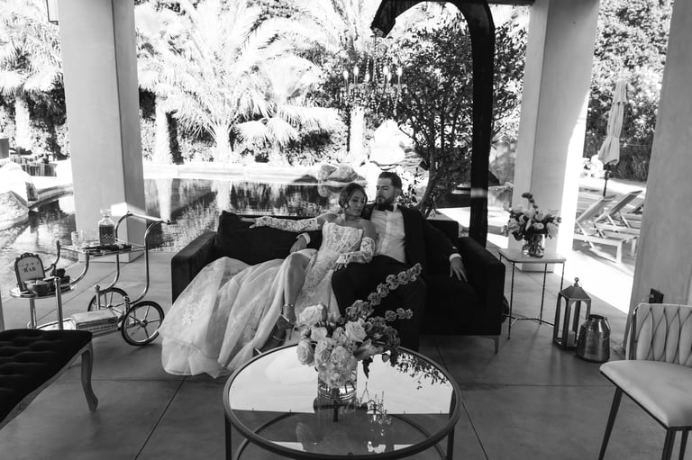 Black and white photo of a bride and groom sitting together in an elegant covered patio with columns, palm trees, and floral arrangements visible