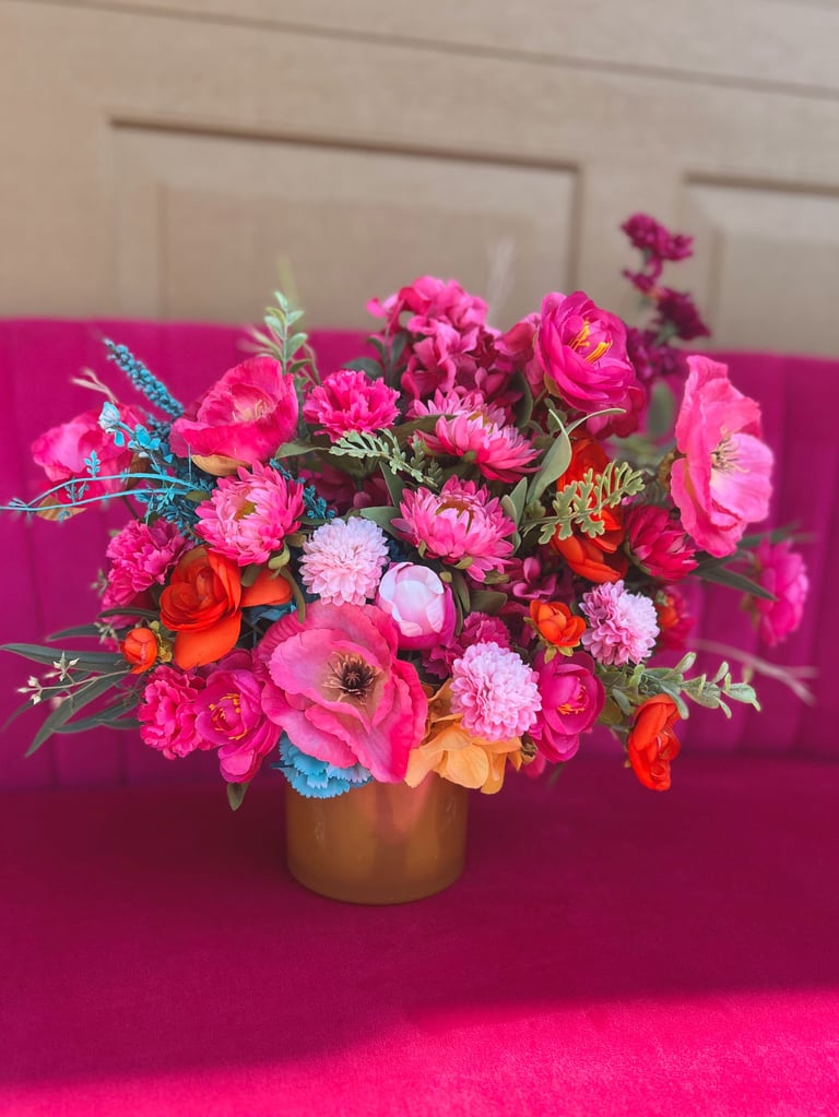 Vibrant bouquet of pink, red, and orange flowers with blue accents in a gold vase on a bright pink surface