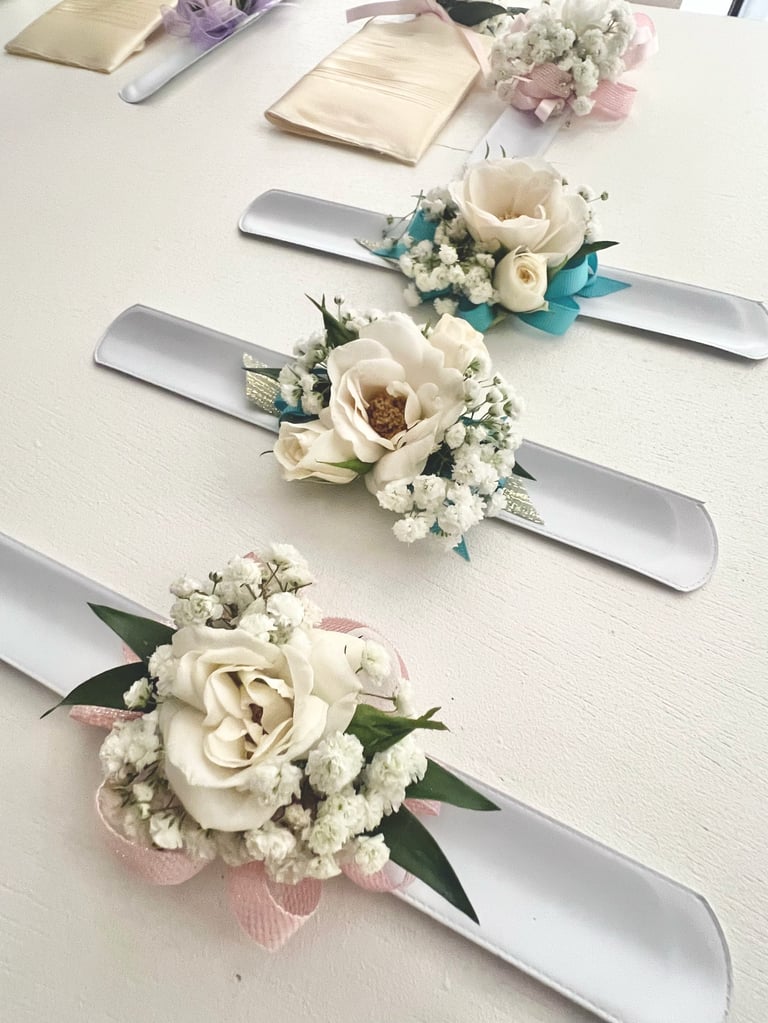 Three bridal corsages with cream roses, babys breath, and ribbon displayed on a white surface with bridesmaid gifts in background