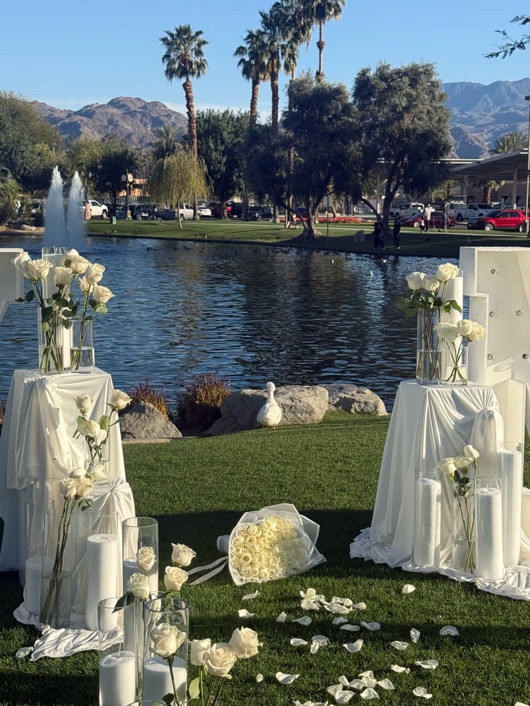 Outdoor wedding ceremony setup by a lake with white draped pedestals, flowers, and mountains visible in the background