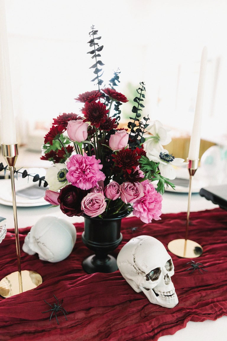 Gothic table centerpiece with dark red and pink flowers in black vase, white skull decoration, and candles on burgundy runner