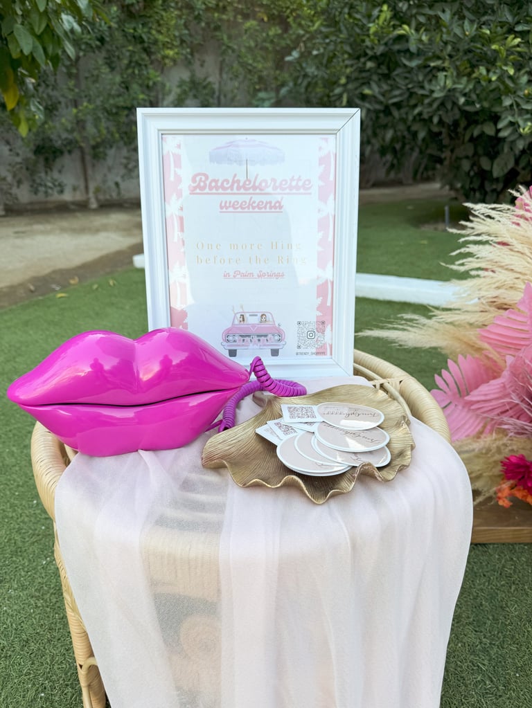 Bachelorette party table with pink lips decoration, welcome sign, and white plates on sheer tablecloth in outdoor garden setting