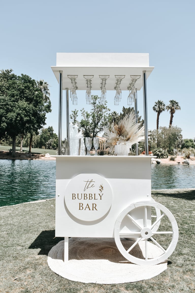 White bubbly bar cart by a lake with palm trees, decorated with hanging flowers and champagne flutes for outdoor events