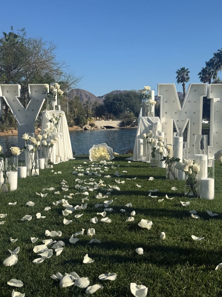 Outdoor wedding ceremony setup with white Y and M letters flanking an aisle covered in white flower petals leading to a waterfront backdrop