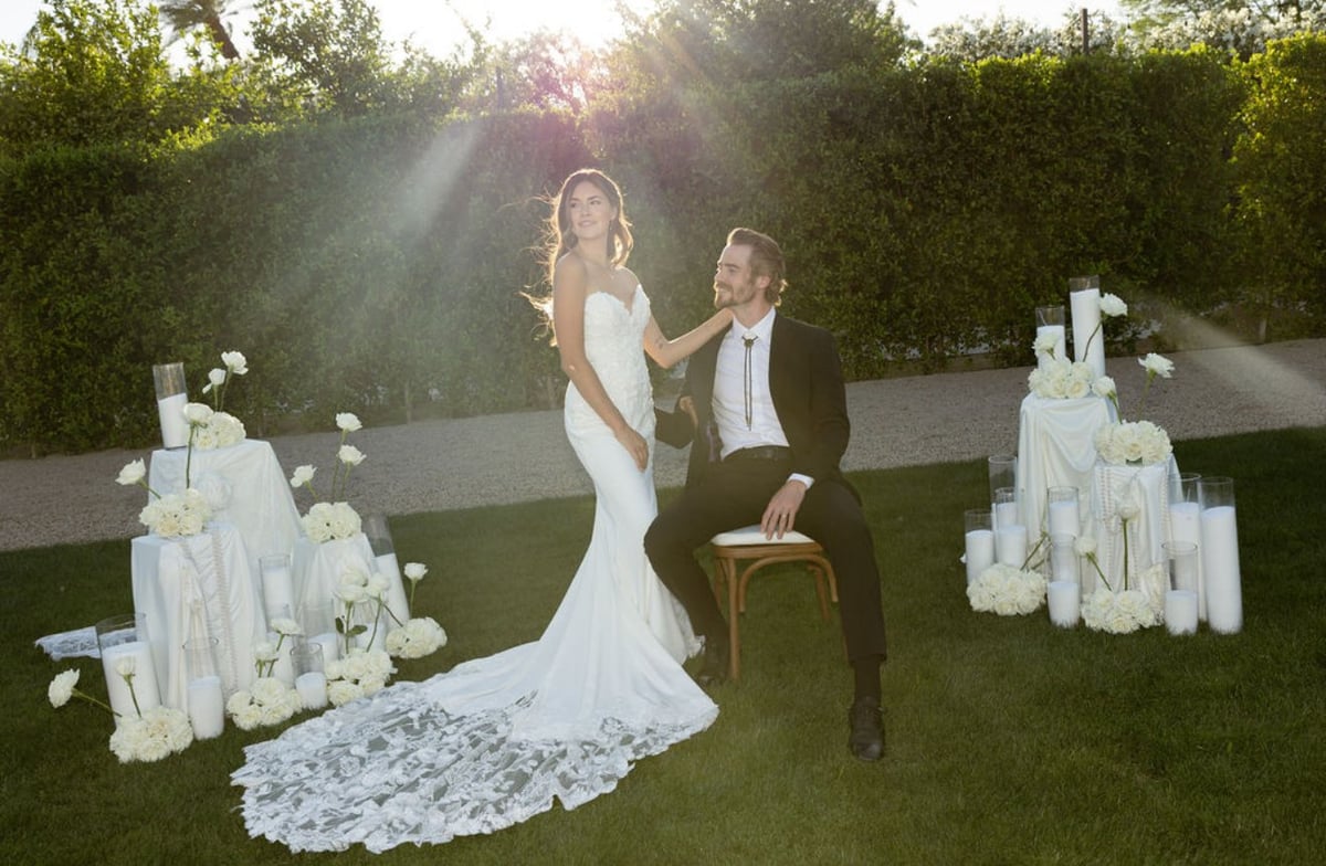 Bride in white lace gown and groom in suit dancing outdoors in garden with floral arrangements at sunset