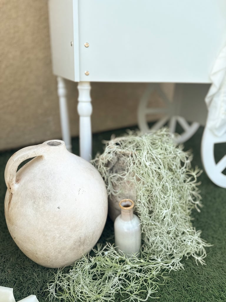 A round cream-colored ceramic vessel with handle next to dried grass, small white bottle, and white furniture on green surface