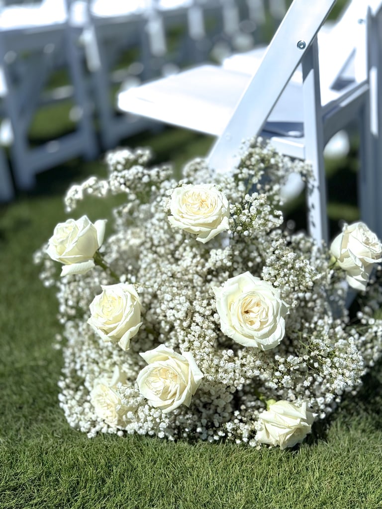 White roses and babys breath flowers arranged at the end of an aisle with white chairs for a wedding ceremony