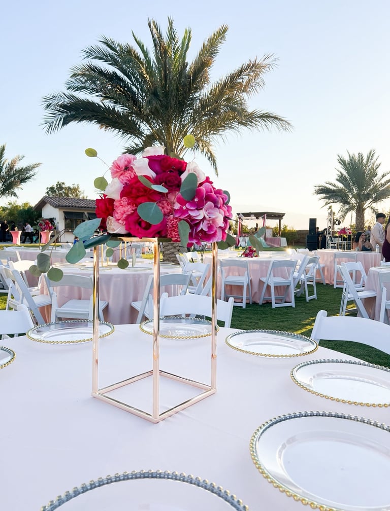 Outdoor wedding reception venue with decorated tables, vibrant pink floral centerpieces, palm trees, and elegant place settings on white linens