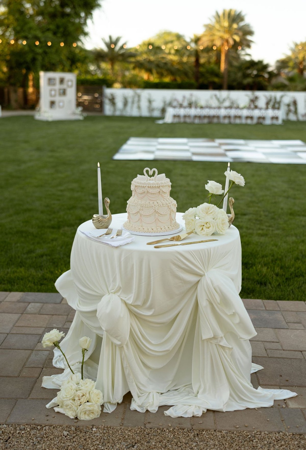 Elegant white wedding cake table with tiered cake, candles, and flowers in an outdoor garden venue with palm trees