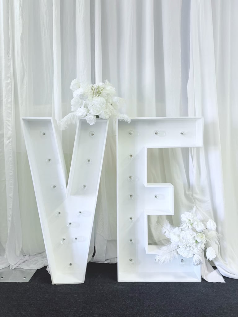 Large white marquee letters V and E decorated with small lights and white flowers against sheer curtains
