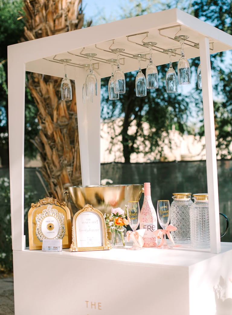 Elegant white outdoor bar cart with hanging glassware, gold accents, champagne bottle, and floral arrangements