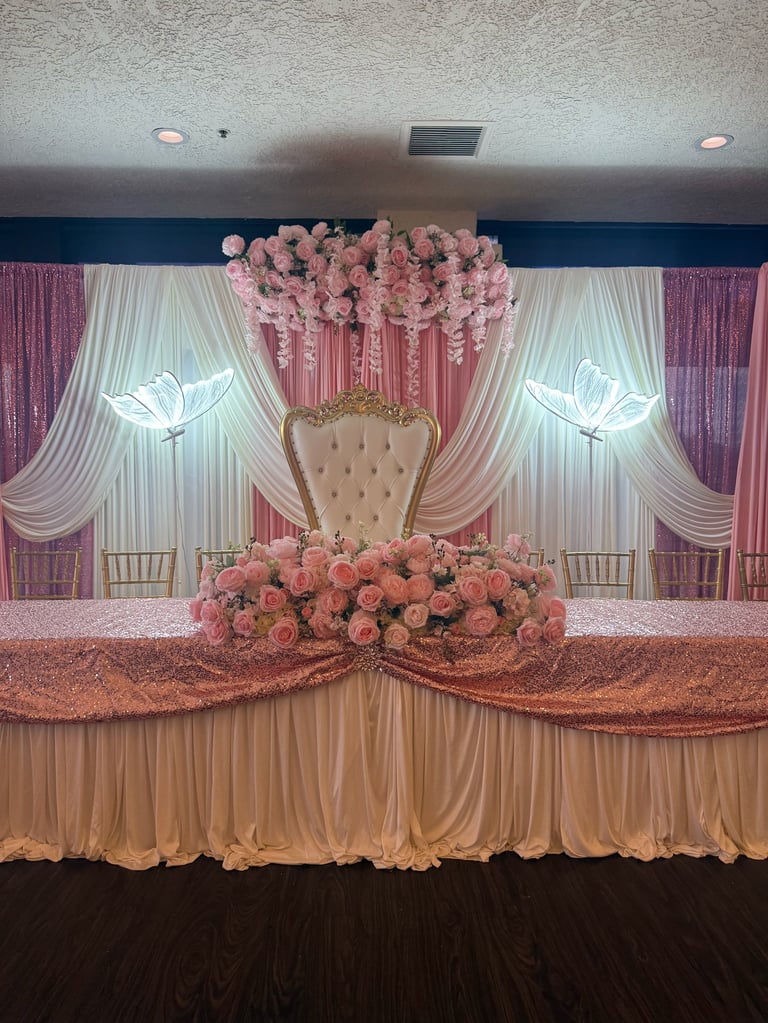 Elegant wedding stage with pink and cream drapes, large floral arrangements, ornate gold chair, and decorative neon wing lights