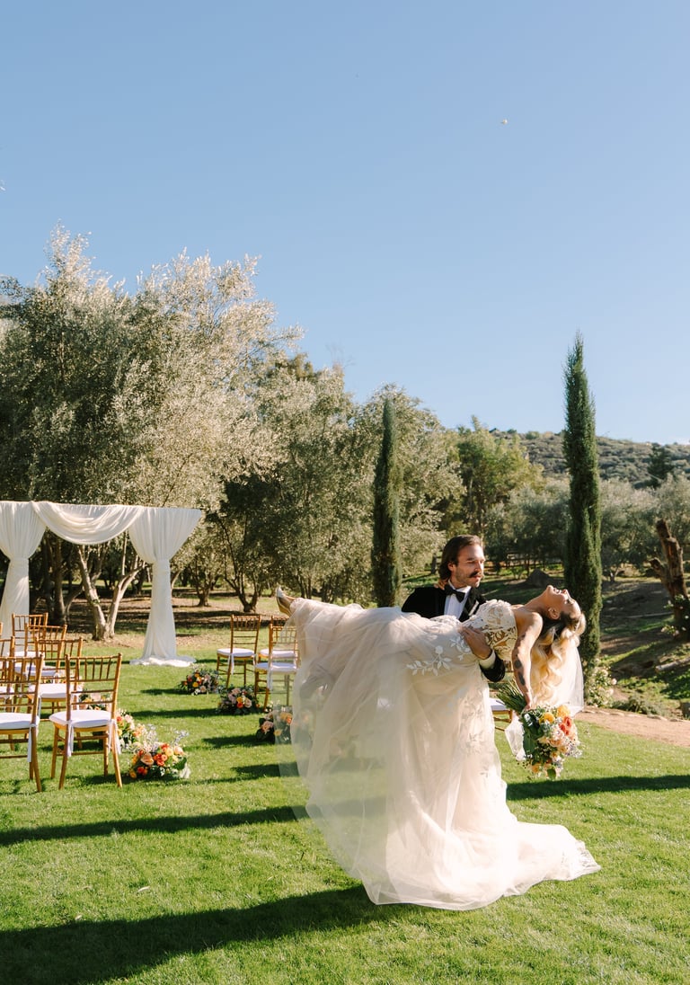 Groom and bride dancing outdoors in a garden ceremony setting with olive trees and draped white fabric visible in the background