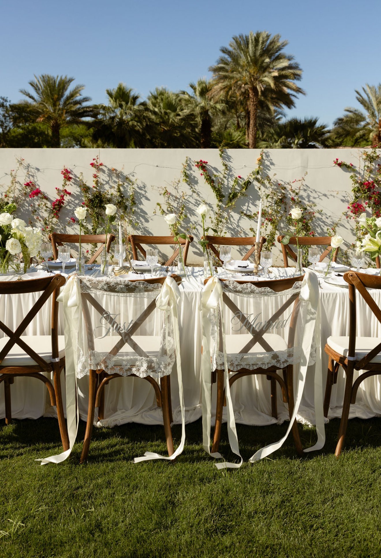 Outdoor wedding reception table with white linens, wooden chairs, and floral arrangements against a white wall and palm trees