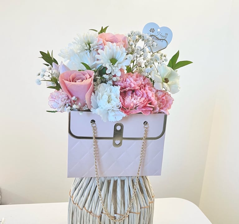 Floral arrangement in purple handbag-shaped vase with pink roses, blue flowers, babys breath, and foliage on white surface