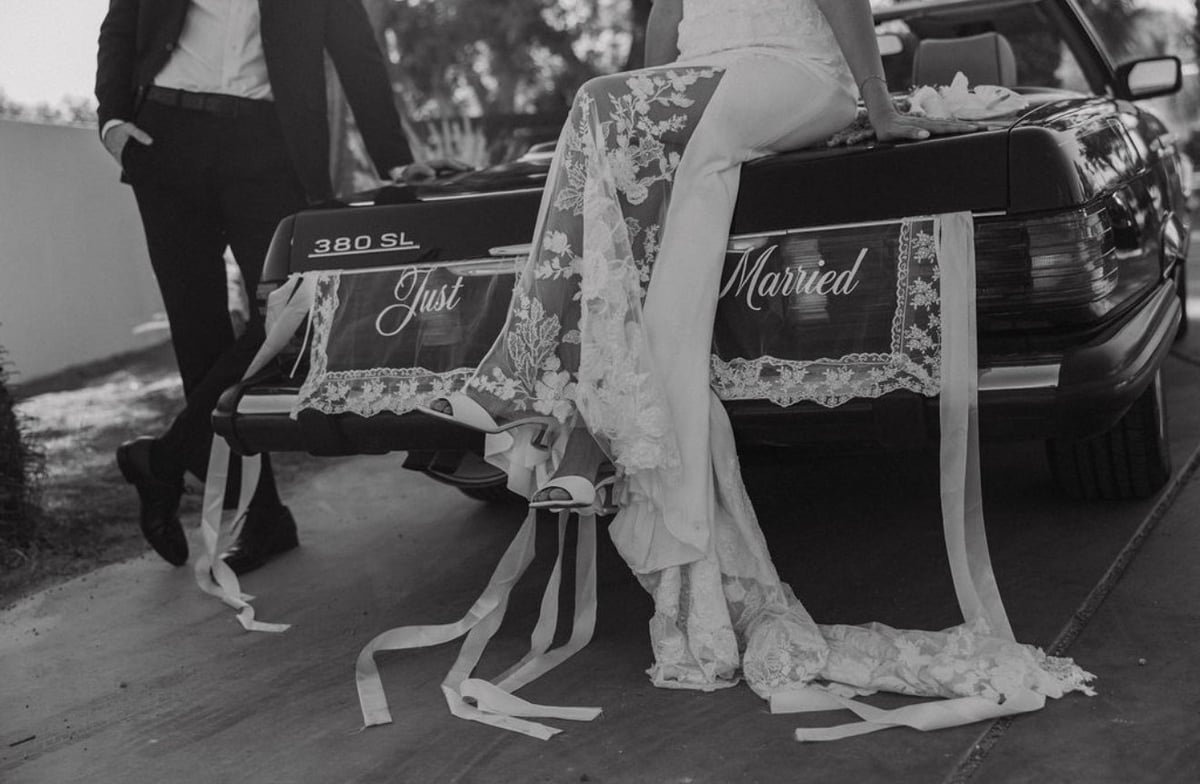 Black and white photo of bride in lace dress with Just Married sign on vintage car with groom