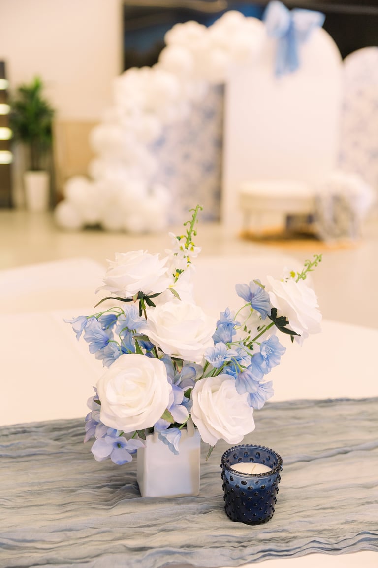White and blue floral arrangement in a white vase next to a dark blue candle on a marble surface
