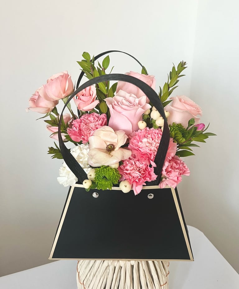 Black handbag-shaped vase with mixed pink roses, carnations, and white flowers with green foliage on white surface