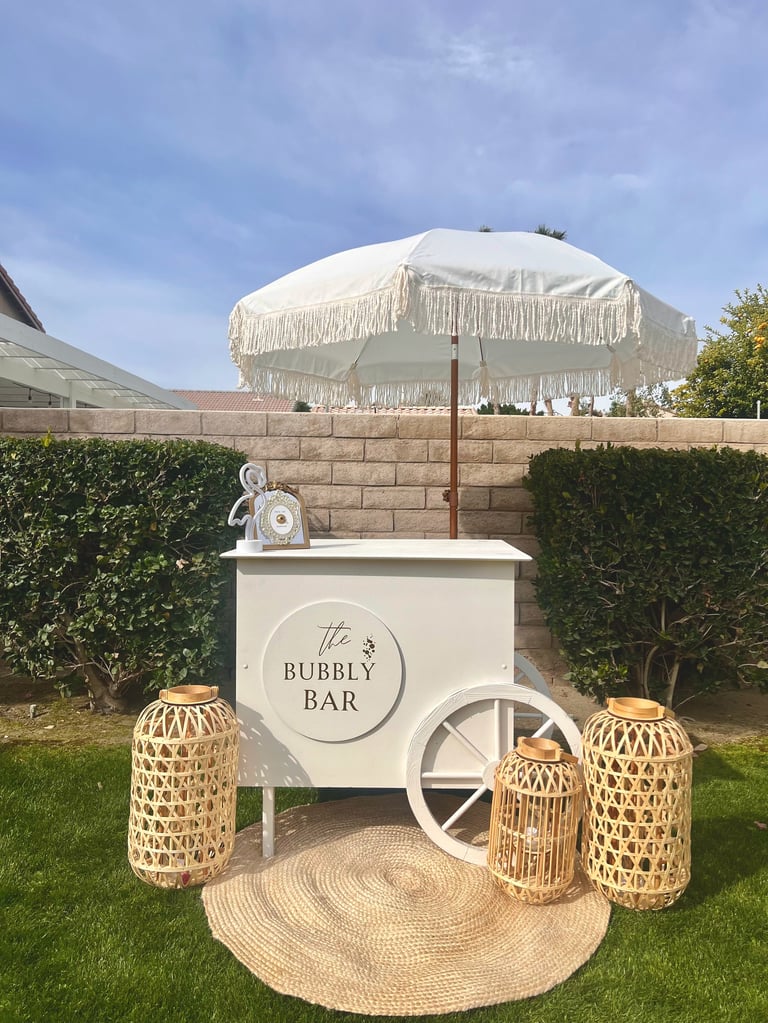 White bubbly bar cart with fringe umbrella on a lawn, decorated with woven lanterns and a circular jute mat
