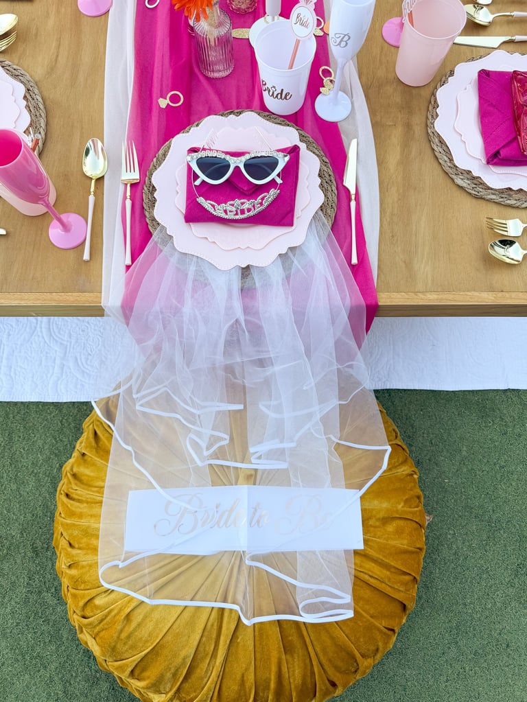 Overhead view of a colorful bridal shower table setting with pink and gold decorations, featuring a place setting with cat-eye sunglasses design and tulle accents.