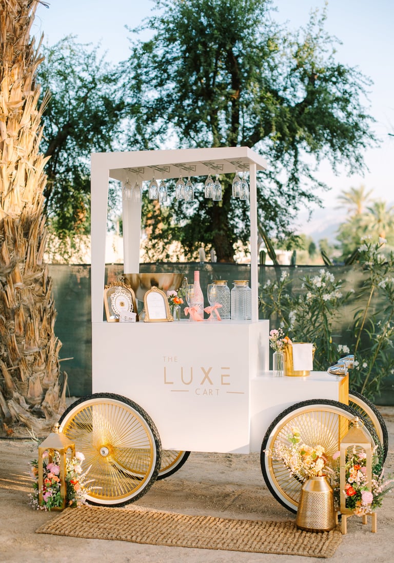 White Luxe Cart vendor stand with gold wheels, flowers, and products displayed in an outdoor garden setting.
