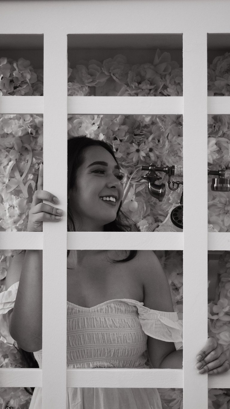 Black and white portrait of a smiling woman in a strapless white top behind a grid window with flowering plants in background