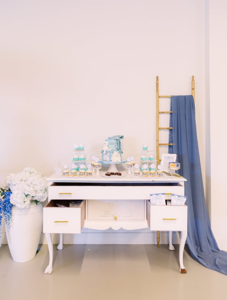 Elegant white dresser styled with blue baby shower displays, flowers in vase, wooden ladder with blue fabric backdrop