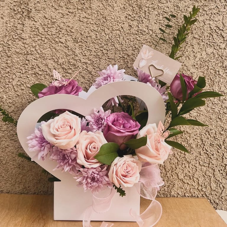 Heart-shaped white box with pink and mauve flowers including roses and carnations on a textured beige background