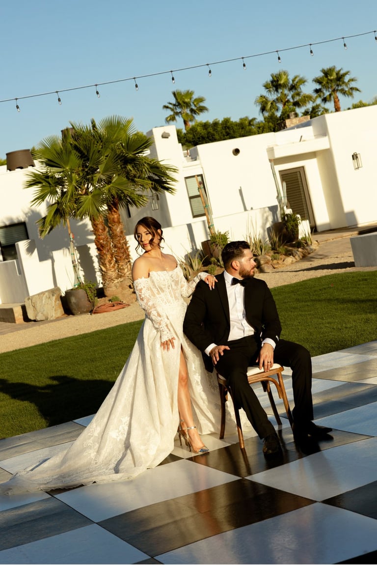 Bride and groom sitting together on a wooden chair in front of a modern white house with palm trees and string lights overhead