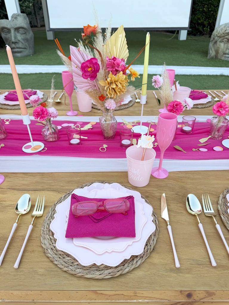 Outdoor table setting with pink and gold theme, featuring place settings with pink napkins, floral centerpiece, and decorative candles
