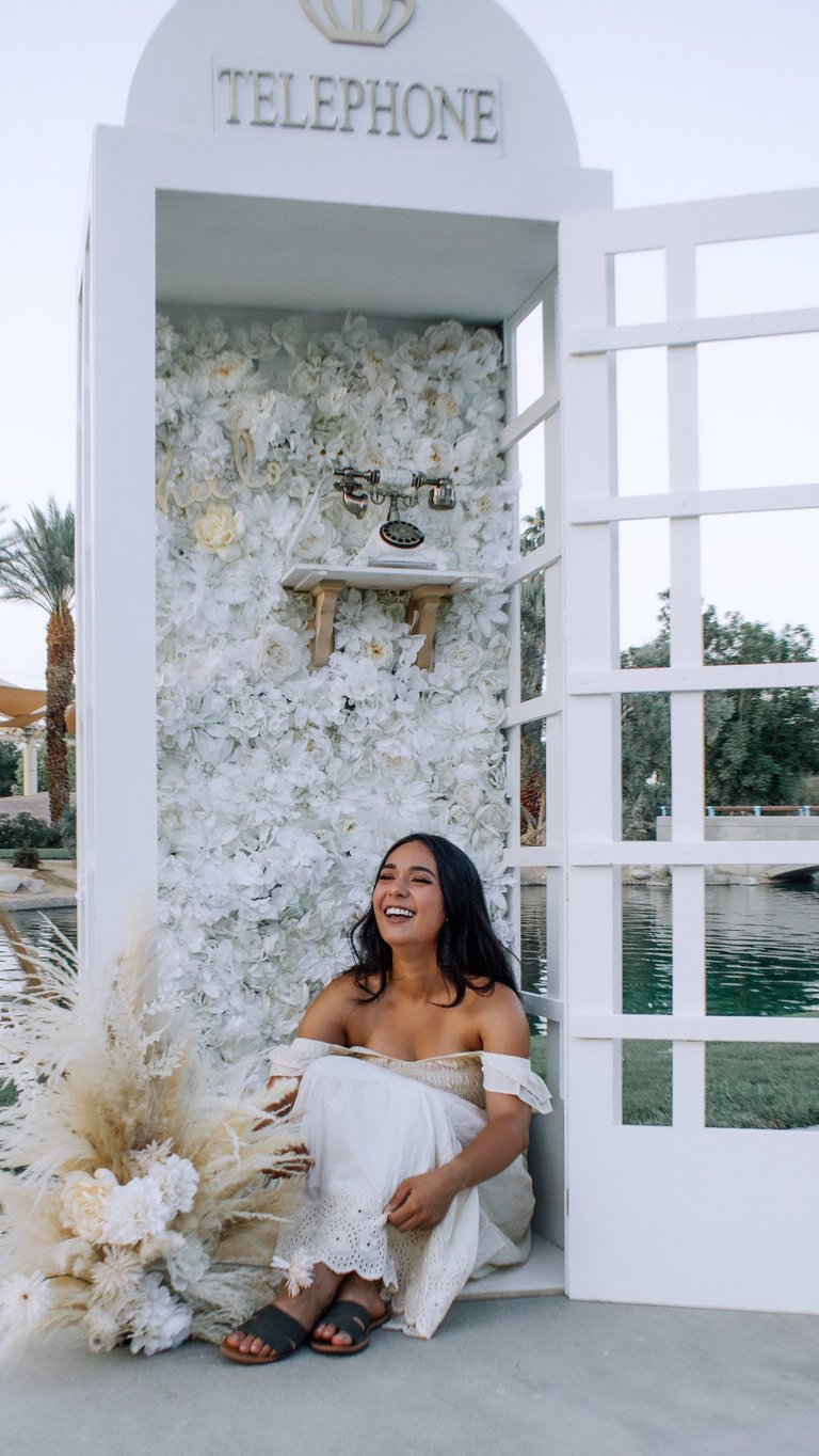 Woman in white dress smiling inside a vintage white telephone booth decorated with white flowers and greenery near water