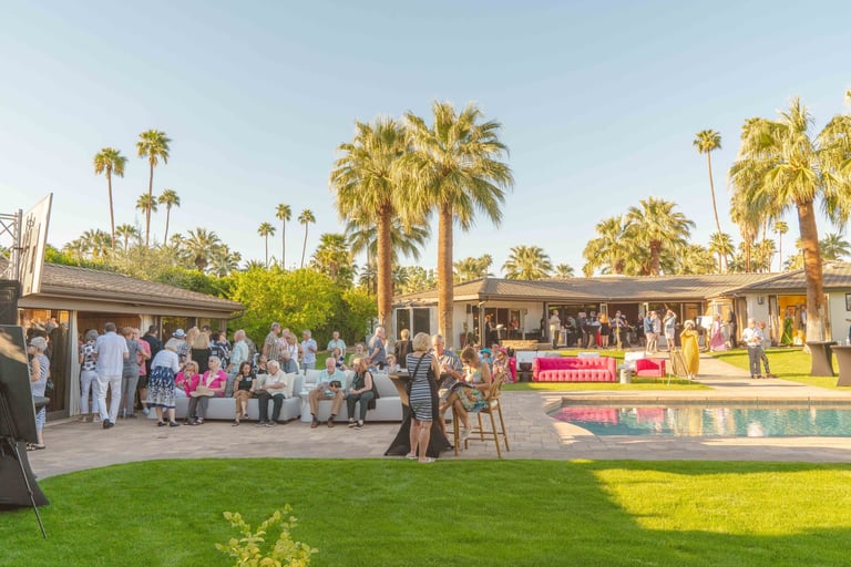 Outdoor event venue with guests on green lawn, pool, palm trees, and modern architectural structures under clear sky