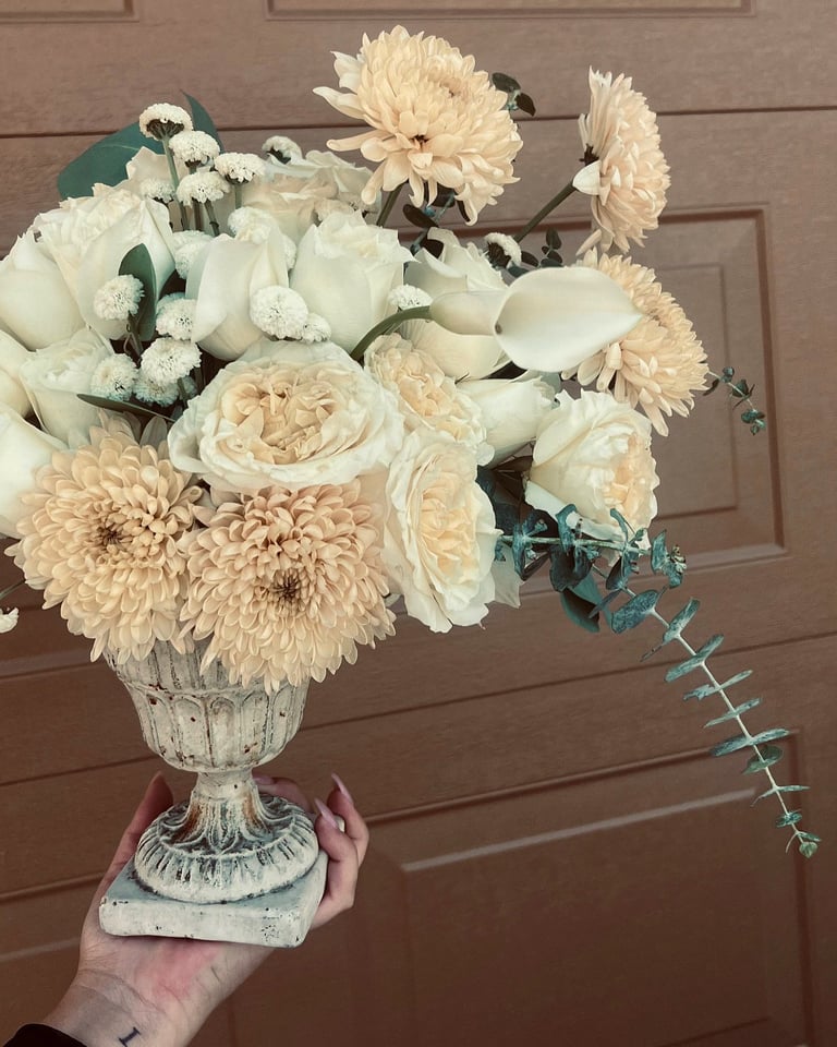 Hand holding a decorative urn vase filled with cream and peach flowers including dahlias, roses, and greenery against a brown wooden door background.