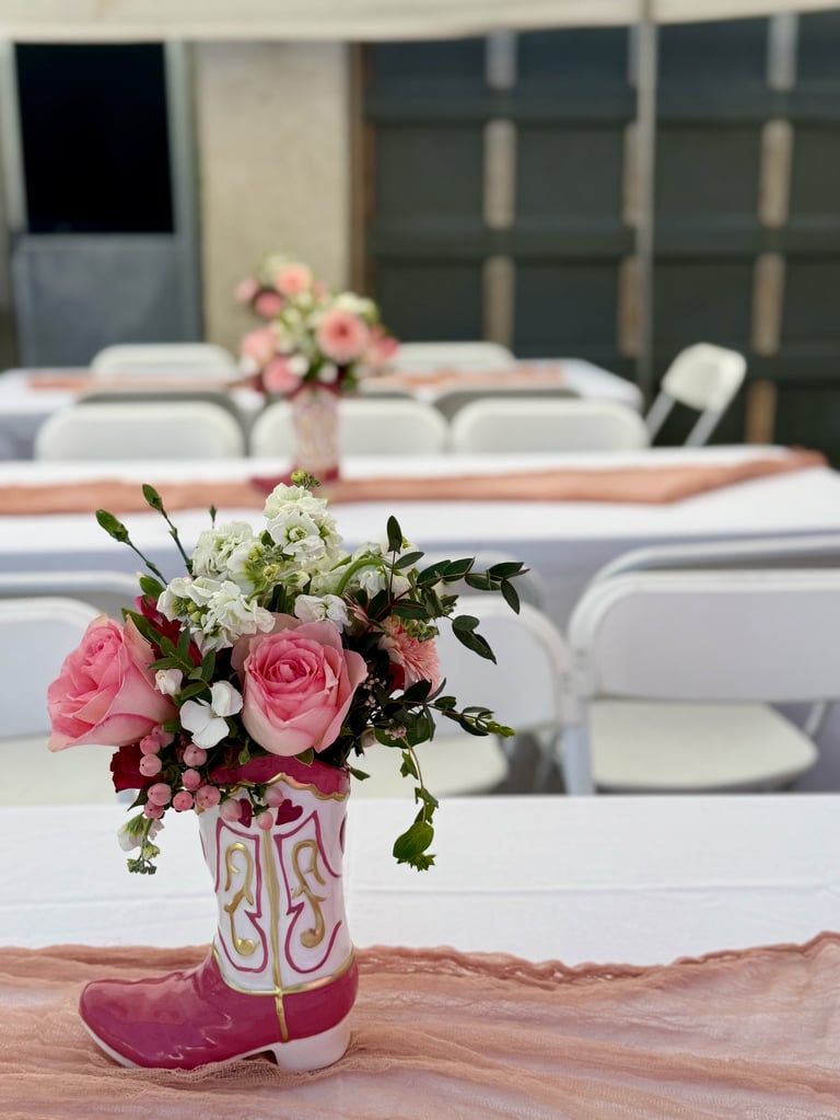 Flower arrangements in decorative boot vases on tables set for an event, with chairs and additional floral displays in the background