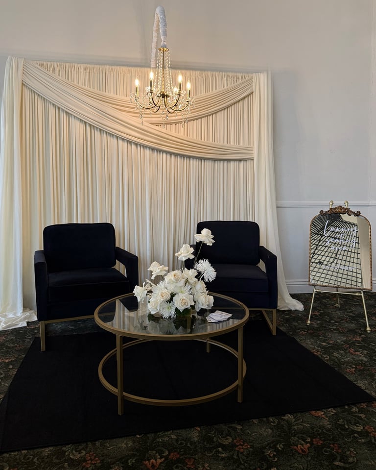 Elegant sitting area with two black velvet armchairs, round glass table with white flowers, and cream draped curtains with chandelier