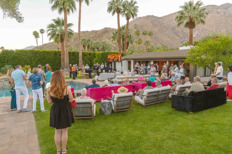 Outdoor event on a manicured lawn with pink-draped seating, palm trees, and mountains in the background