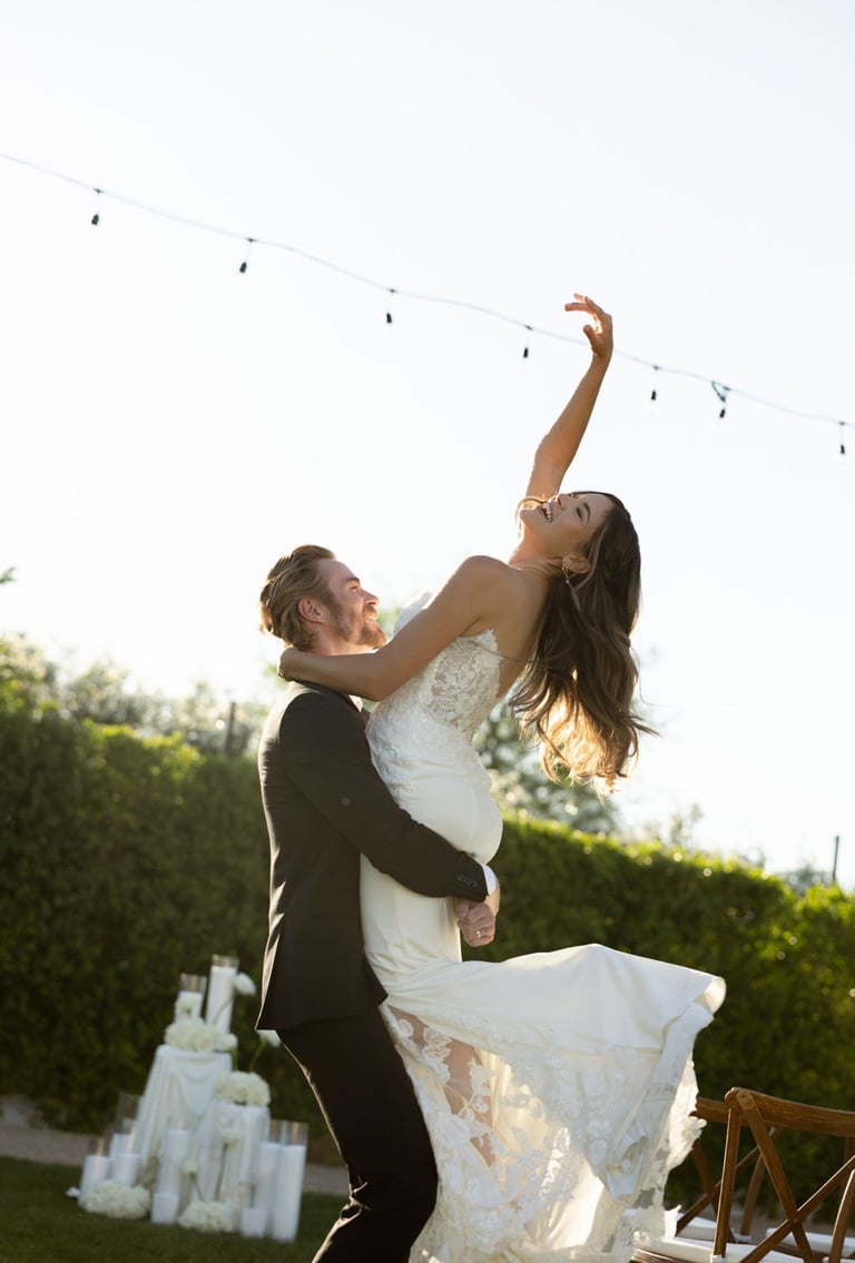 Groom lifting bride in white wedding dress outdoors with string lights overhead