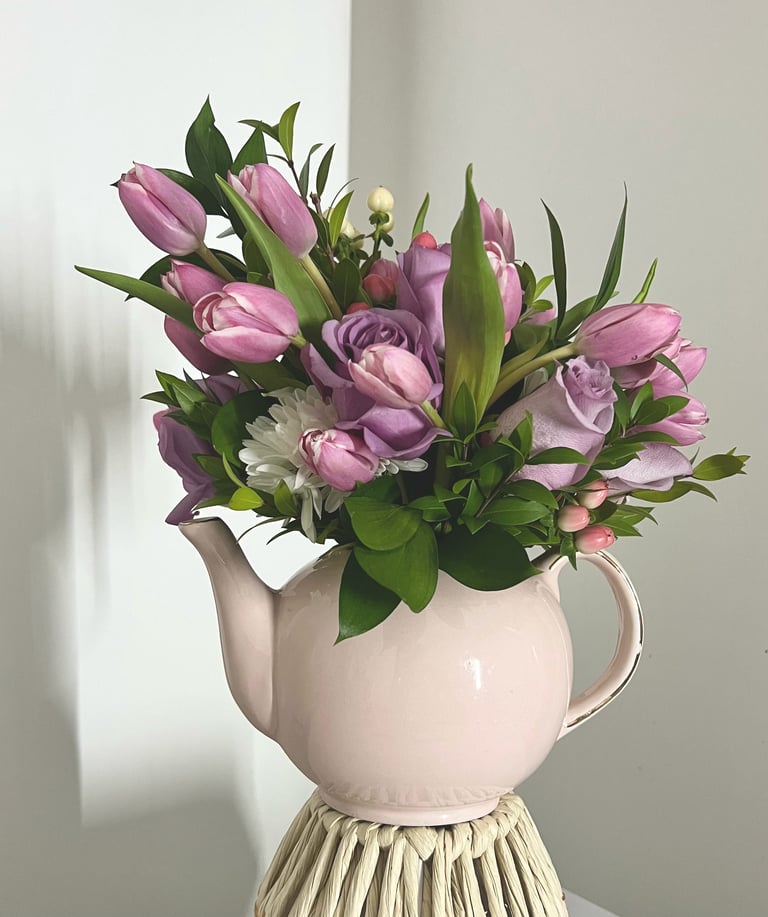 Bouquet of pink and purple tulips with green foliage arranged in a white ceramic pitcher vase.