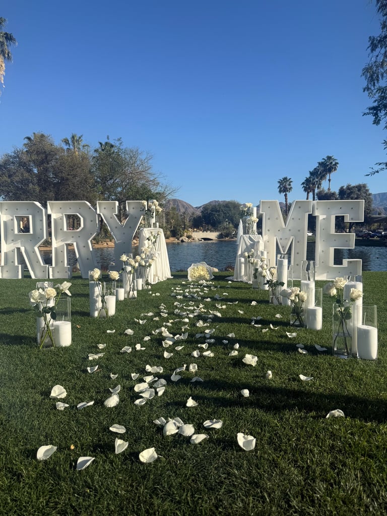 Wedding ceremony setup with large white MARRY ME letter decorations, white flowers, and white chairs along a waterfront lawn with palm trees and mountains in background