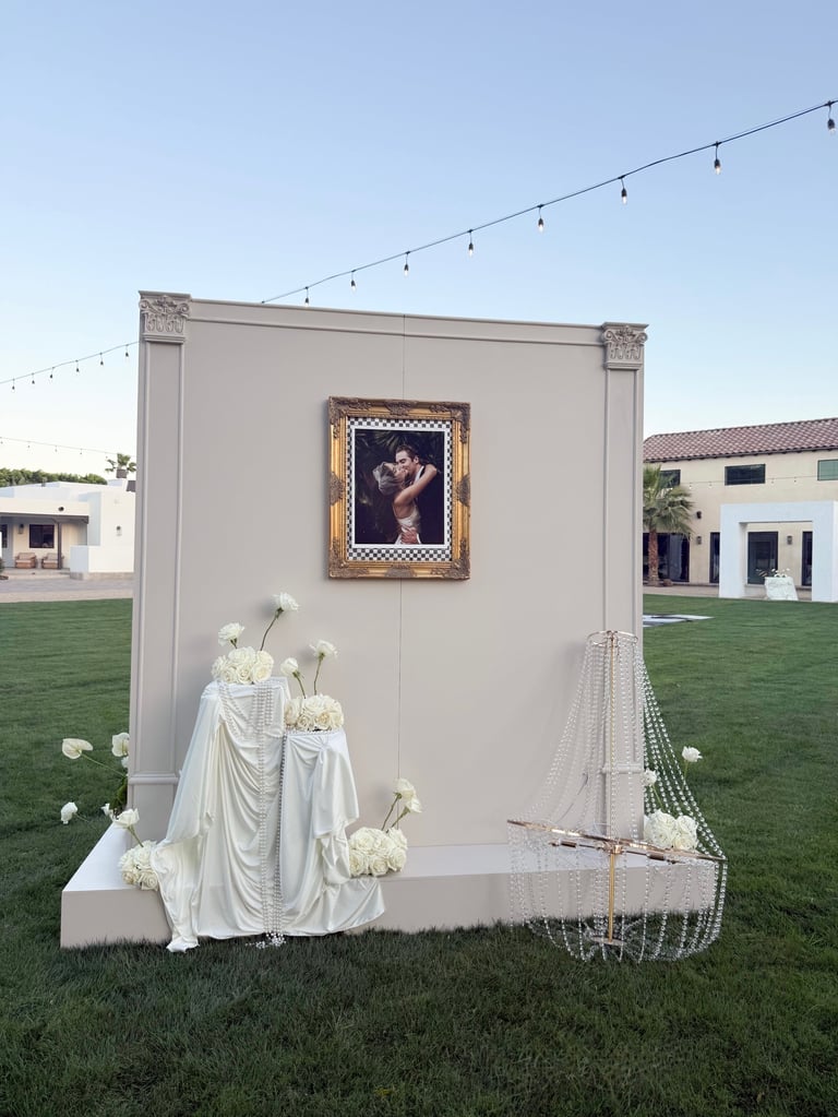 Outdoor wedding photo booth with cream wall, framed portrait, white fabric draped figures, white flowers, and fishing net decoration in grassy garden setting