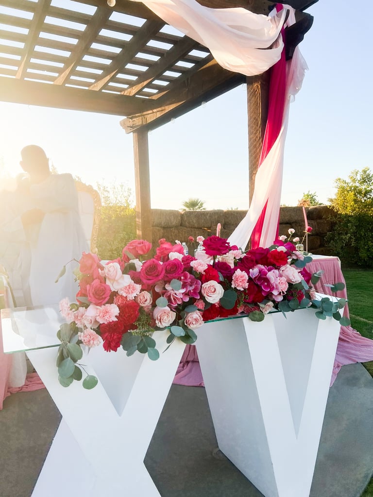 Outdoor wedding altar decorated with pink, red, and white flowers in white vases under a wooden pergola with flowing pink fabric