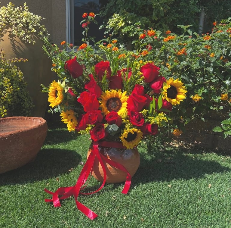 Vibrant floral arrangement with red roses and yellow sunflowers in terra cotta vase tied with red ribbon, displayed on lawn