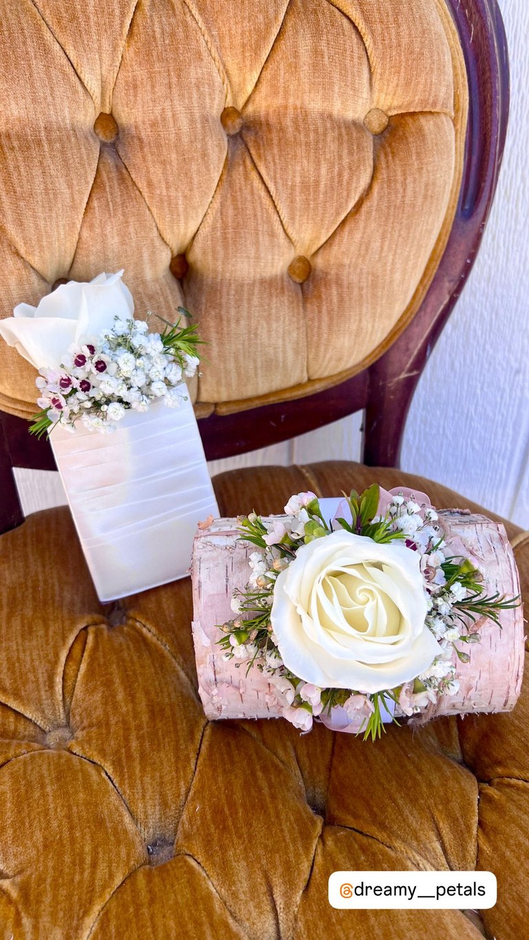 Two floral arrangements on a tufted beige headboard: a pink corsage with white rose and babys breath, and a white flower arrangement in a frosted vase