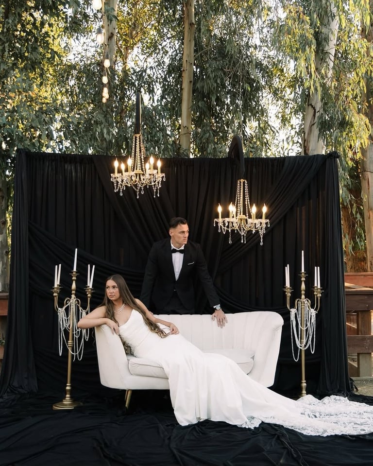 Bride and groom portrait on white sofa with black drapes, chandeliers, candelabras, and tall trees in background