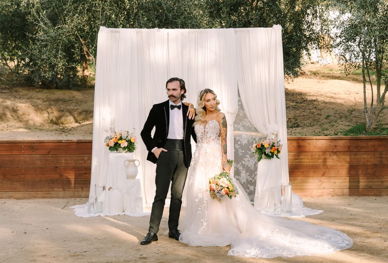 Bride and groom standing together at outdoor wedding ceremony with white draped backdrop and floral arrangements