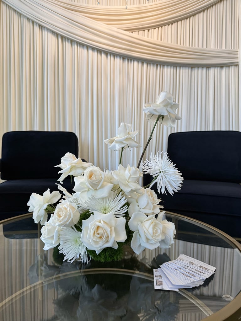 Elegant white floral arrangement on glass table between two black chairs with draped cream curtains in background