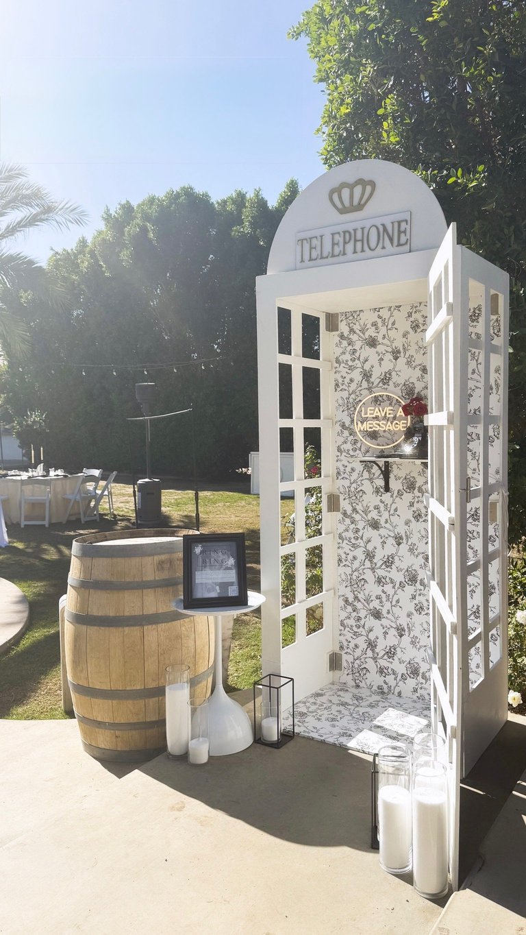 Vintage-style white telephone booth with crown logo at outdoor garden event, featuring wooden barrel and floral wallpaper details