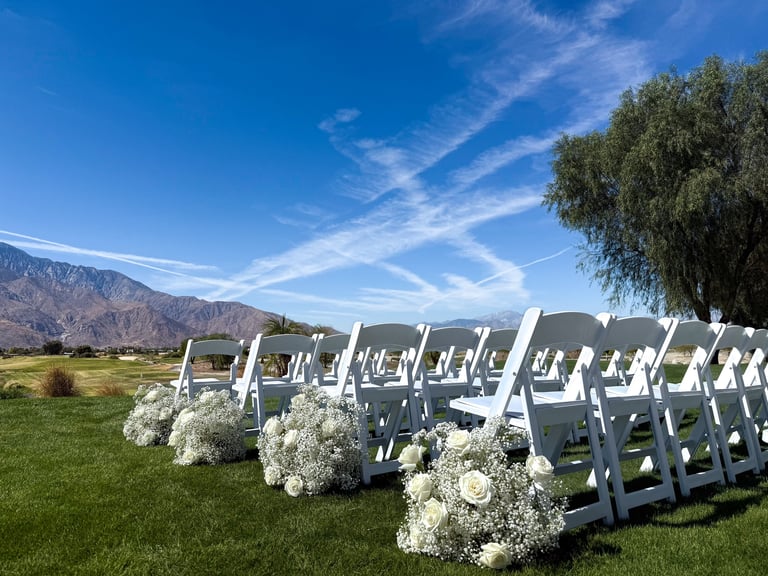 Outdoor wedding ceremony setup with white chairs lined on green lawn, decorated with white floral arrangements and mountain backdrop