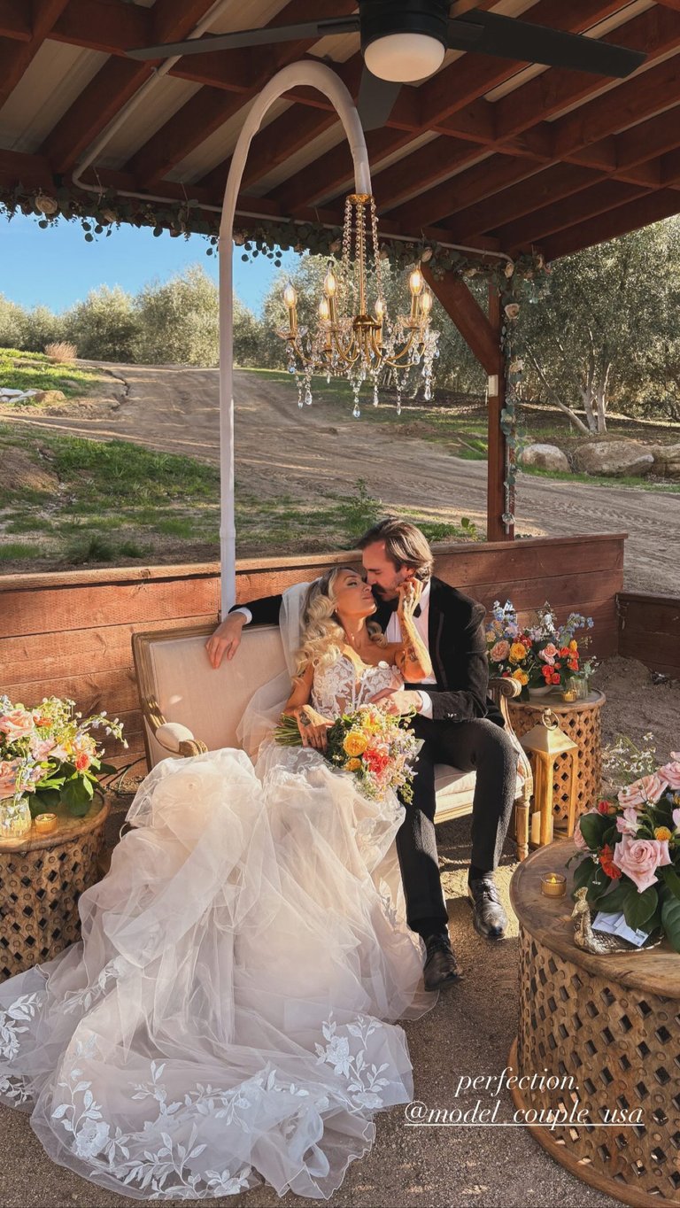 Bride and groom kissing under a rustic gazebo with chandelier, surrounded by floral arrangements and olive trees